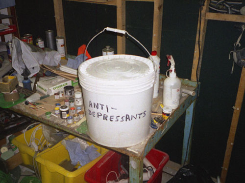 A very messy workbench sits in an equally cluttered room with an unfinished wall. Art supplies are scattered everywhere. In the center is a white plastic bucket which someone has used a marker to label "ANTI-DEPRESSANTS."