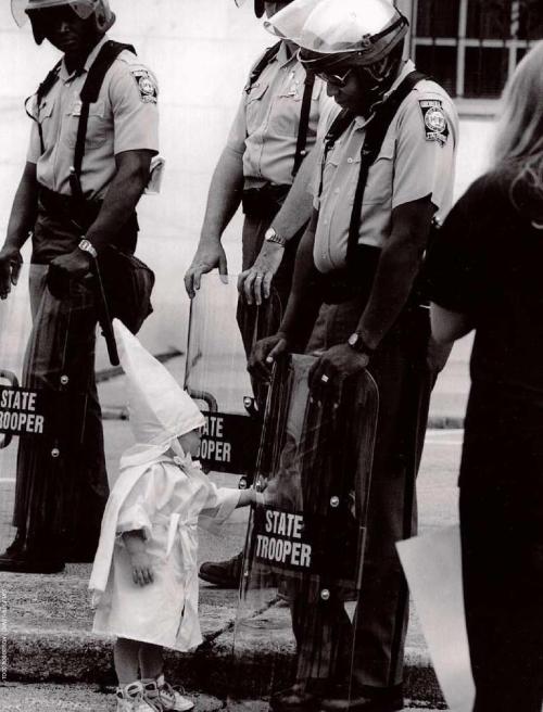 
Here is a Georgia State Trooper in riot gear at a KKK protest in a north Georgia city back in the 80s. The Trooper is black. Standing in front of him and touching his shield is a curious little boy dressed in a Klan hood and robe. I have stared at this picture and wondered what must have been going through that Trooper’s mind. Before the Trooper is an innocent child who is being taught to hate him because of the color of his skin. The child doesn’t understand what he is being taught, and at this point he doesn’t seem to care. Like any other child his curiosity takes hold and he wants to explore this new thing that this man is holding probably because he can see his reflection in it and that’s a neat thing and he wants to check it out. In this picture I see innocence mixed with hate, the irony of a black man protecting the right of white people to assemble in protest against him, temperance in the face of ignorance, and hope that racism can be broken because this young boy may remember that a black man smiled at him once and he didn’t seem so bad after all.
(Picture source)(Paragraph source)

Wow. 