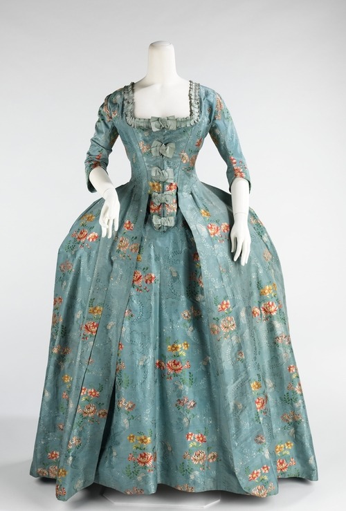 Robe à la Française | 1760-1770 Femmes avec des airs coquets ont été imposante en robes à la française et des robes à l'anglaise pendant toute la période entre 1720 et 1780.  La robe à la française a été dérivée de la robe sacque déshabillé lâche de la première partie du siècle, qui a été plissée à partir des épaules à l'avant à l'arrière.  La silhouette, composée d'un buste en forme d'entonnoir d'alimentation dans de larges jupes rectangulaire, a été inspiré par des conceptions espagnole du siècle précédent et a permis pour des montants expansive du textile avec la décoration délicate curviligne rococo.  Les jupes larges, qui ont souvent été ouverte à l'avant pour exposer un jupon très décorées, ont été soutenus par sacoches créés à partir de rembourrage et des cerceaux de différentes matières telles que la canne, à fanons ou en métal.  Les robes à la française sont réputées pour la beauté de leurs textiles, la coupe des plis creux et de décorations de retour employant jupe, connu sous le nom robings, qui a montré l'imagination infinie et de la variété.