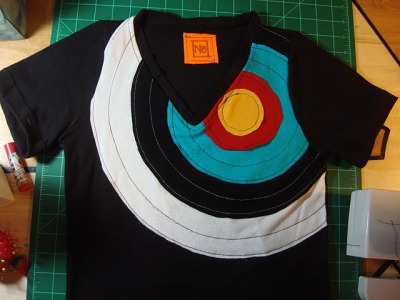 Just finished this new tshirt! Made completely from scratch from recycled thrift-store tshirts, based on my own pattern. I literally just harvested the fabric and constructed an entirely new shirt, vs my first attempt, which used a pre-made tshirt as the base. Do you like archery targets? Me too. I thought it would make a nice inspiration for this design.
I really upped the anti on quality with this version, taking care to reinforce every seam, and include a finished v-neck, which was challenging but I think makes the shirt look much more professional.

Sewing the target onto the shirt, and here are some sketches for future ideas.

It fits!

Trying out a label idea.